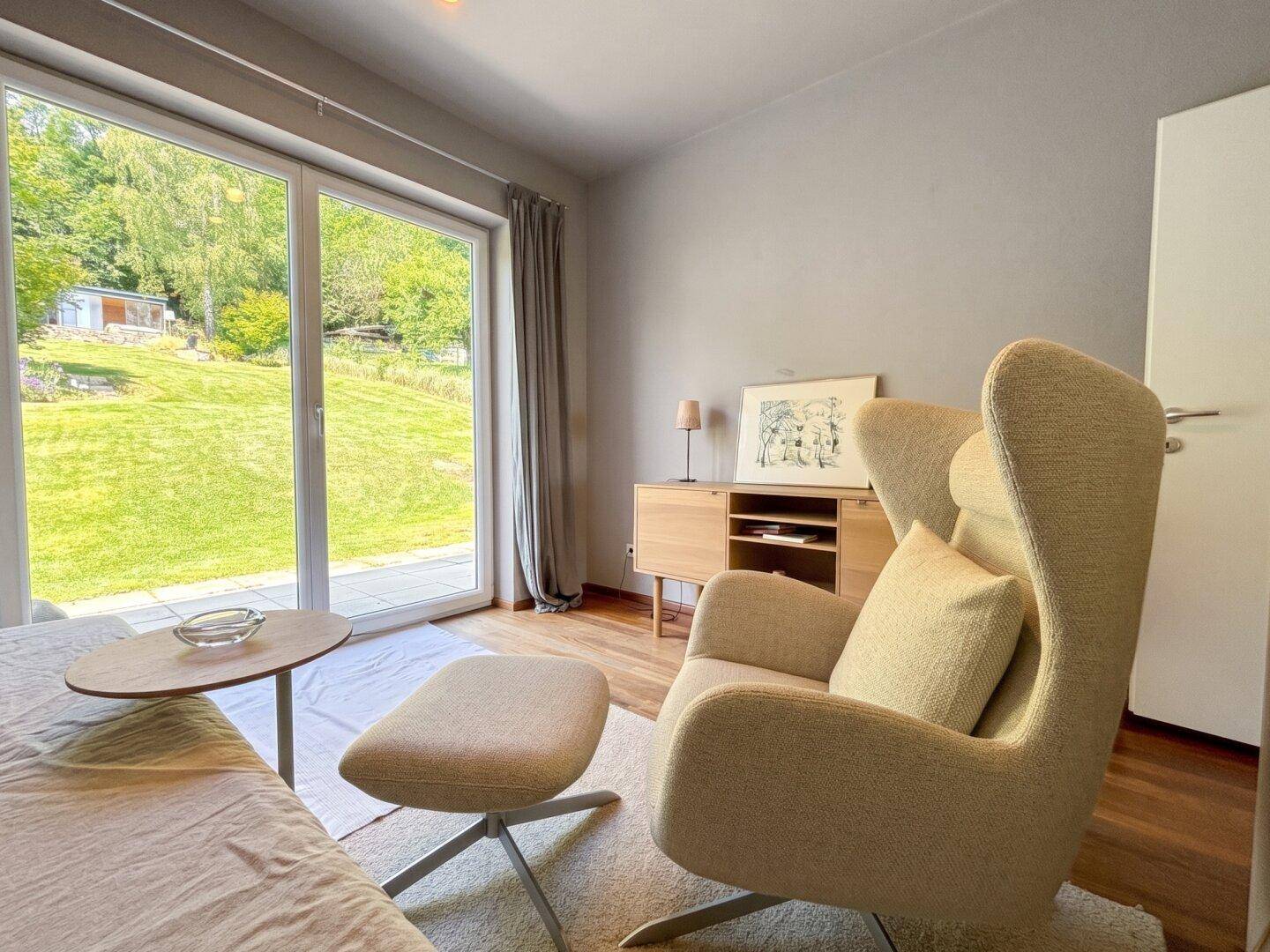 Gemütlicher Raum mit Sessel, Holzboden und großen Fenstern mit Blick auf die grüne Landschaft.