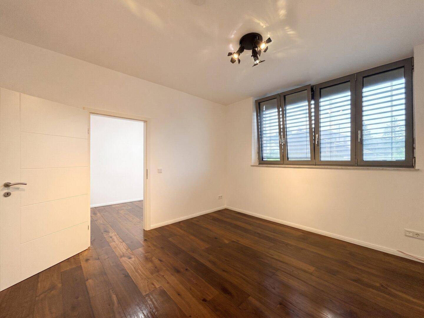 Leeres Zimmer mit dunklem Holzboden und Fenstern mit Jalousien, ideal als Büro oder Schlafzimmer.