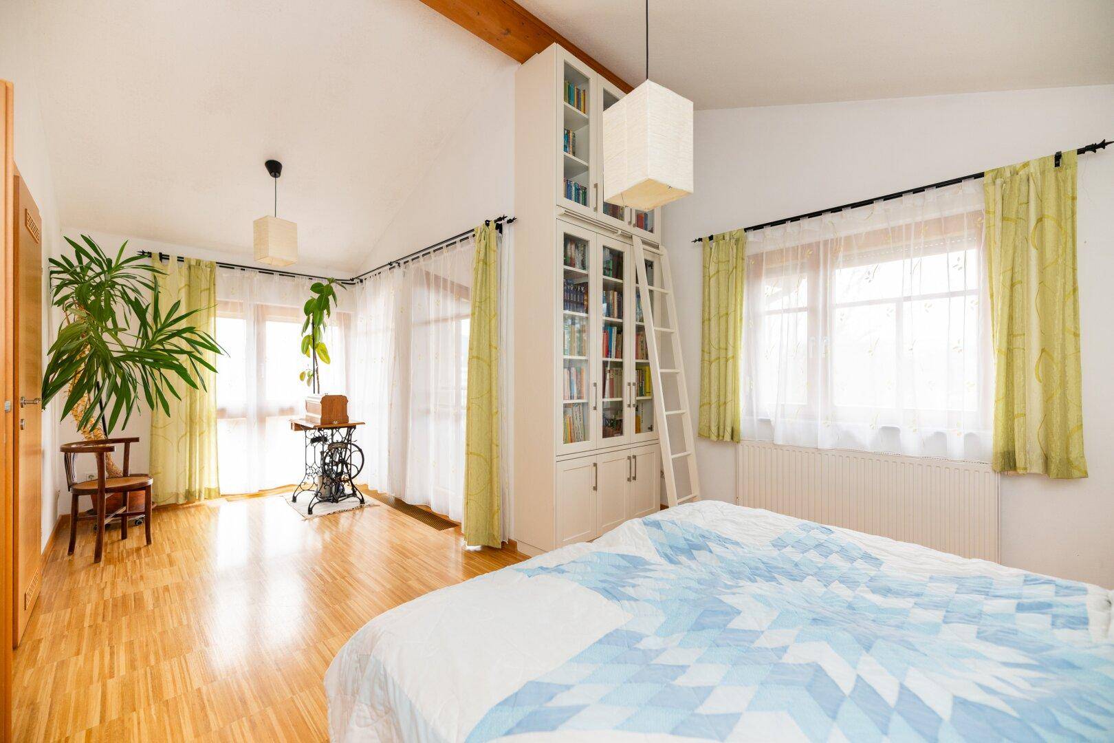 Geräumiges Schlafzimmer mit Holzbalken, hohen Decken, Bücherregal und großen Fenstern.