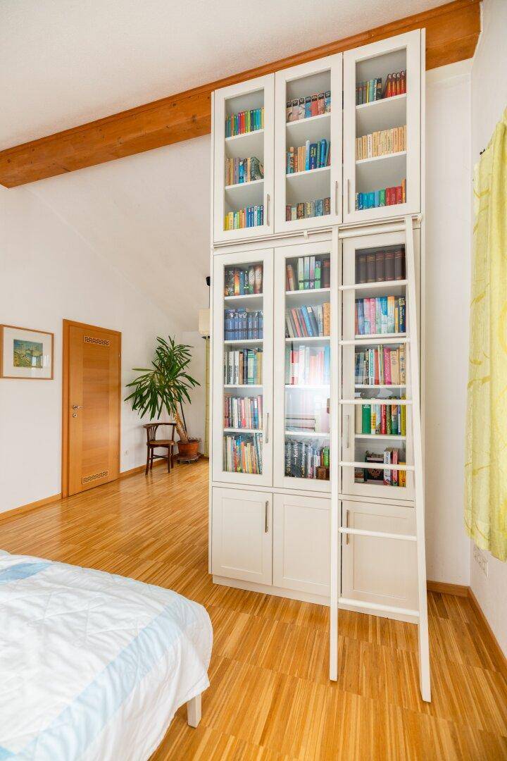 Hohes Bücherregal mit Leiter in einem hellen Schlafzimmer mit Holzboden und Balken.