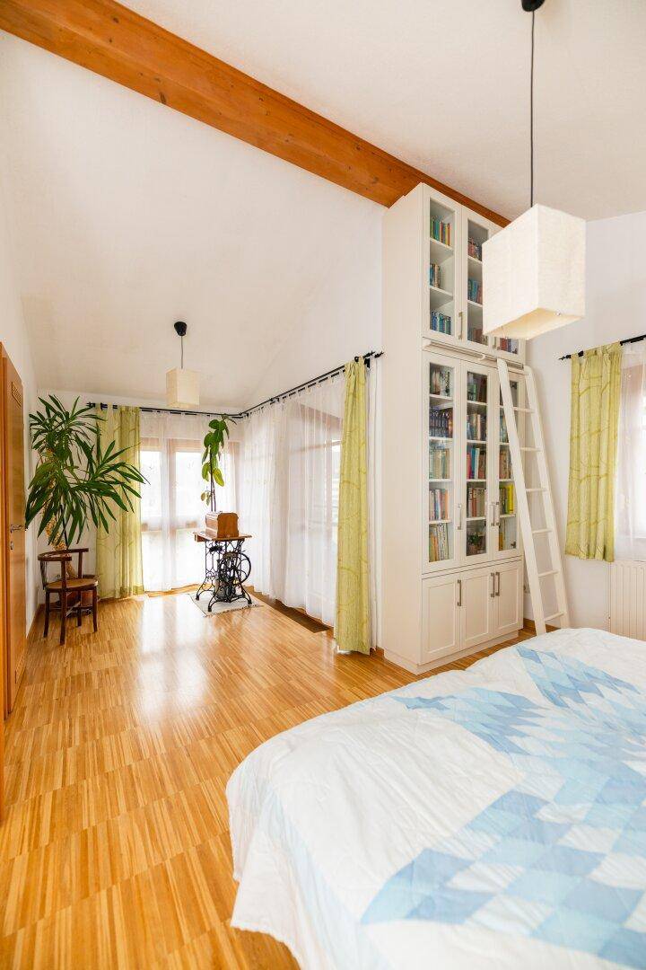 Geräumiges Schlafzimmer mit Holzbalken, hohen Decken, Bücherregal und großen Fenstern.