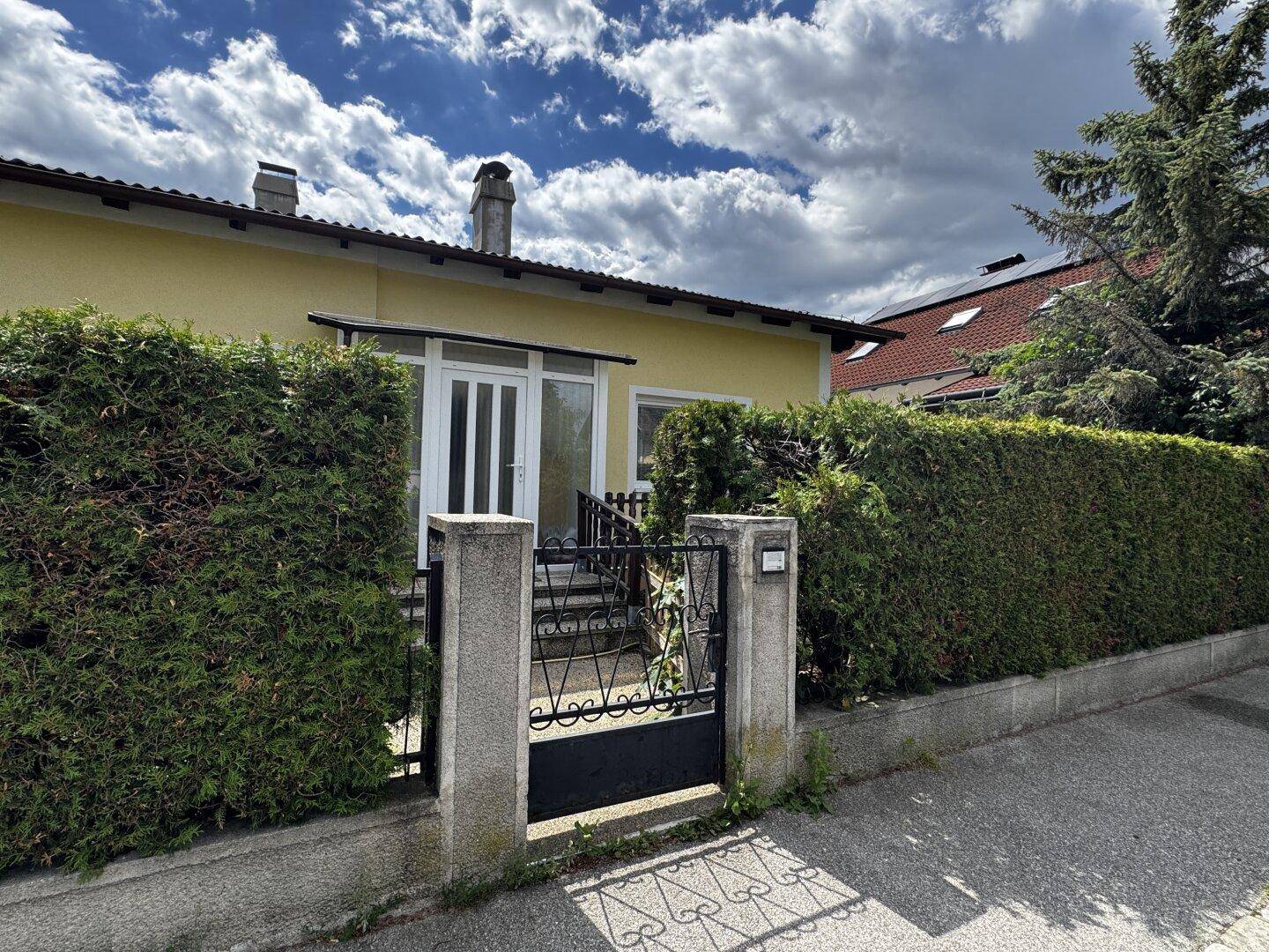 Vorderansicht eines Einfamilienhauses mit hellgelber Fassade, Eingangsbereich und grüner Hecke zur Straße.