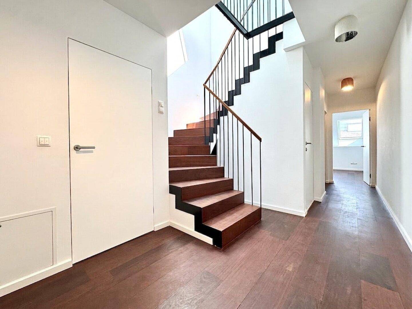 Heller Flur mit eleganter Holztreppe und dunklem Holzboden, der zu den oberen Etagen führt.