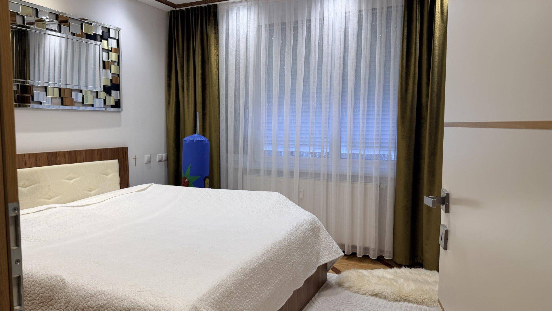 Schlafzimmer mit großem Bett, Holzboden, dekorativem Spiegel und Vorhängen am Fenster.