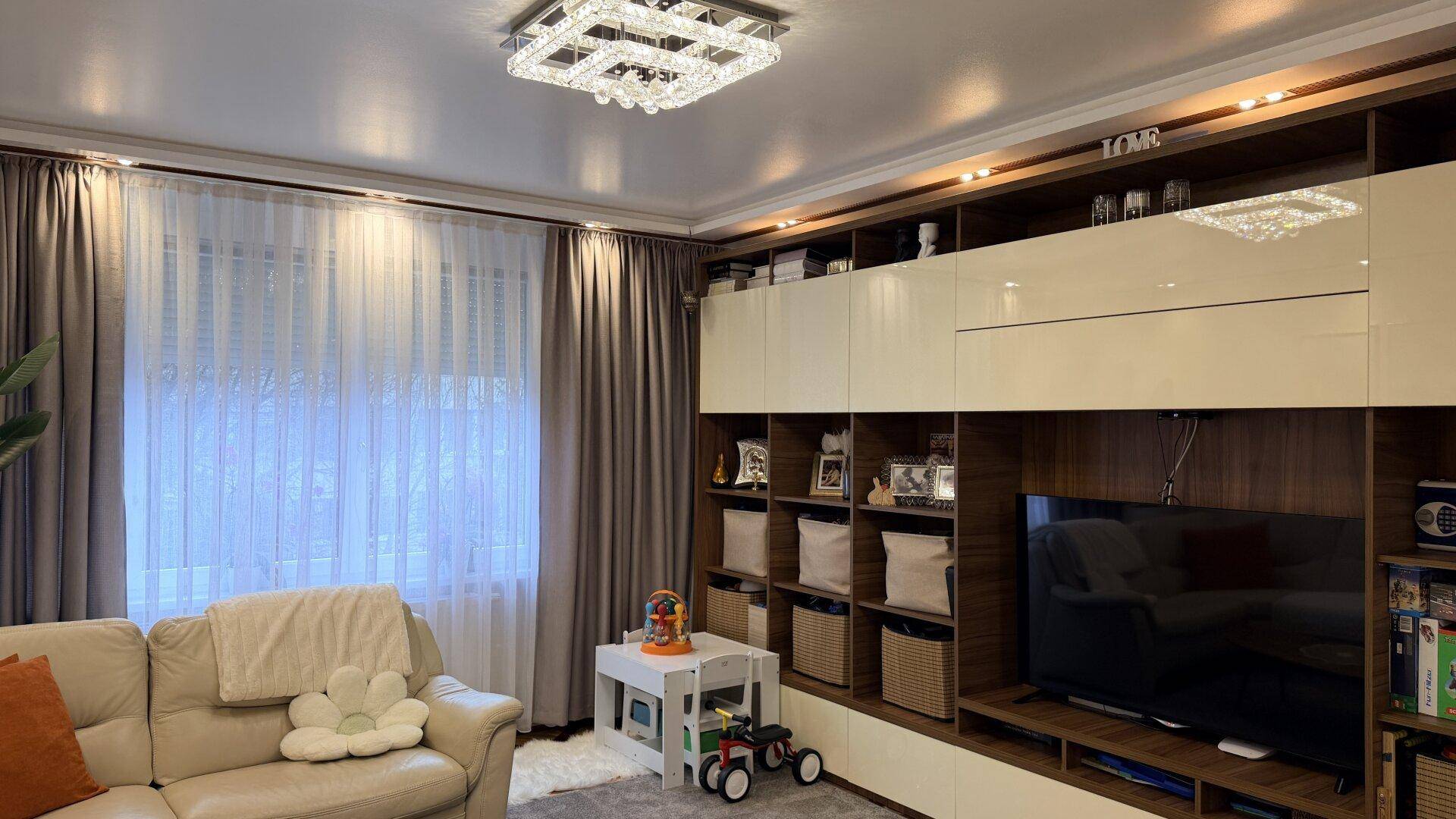 Modern eingerichtetes Wohnzimmer mit großem Ecksofa, TV-Möbeln und einem Fenster mit Vorhängen.