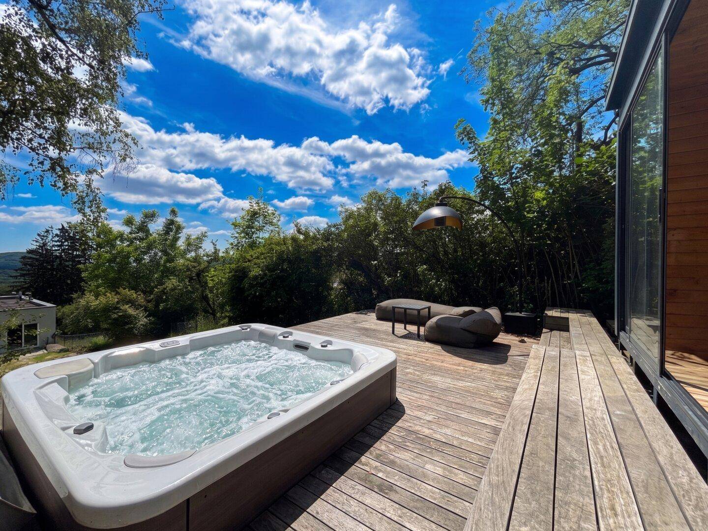 Holzterrasse mit sprudelndem Whirlpool, umgeben von üppiger Vegetation unter blauem Himmel.
