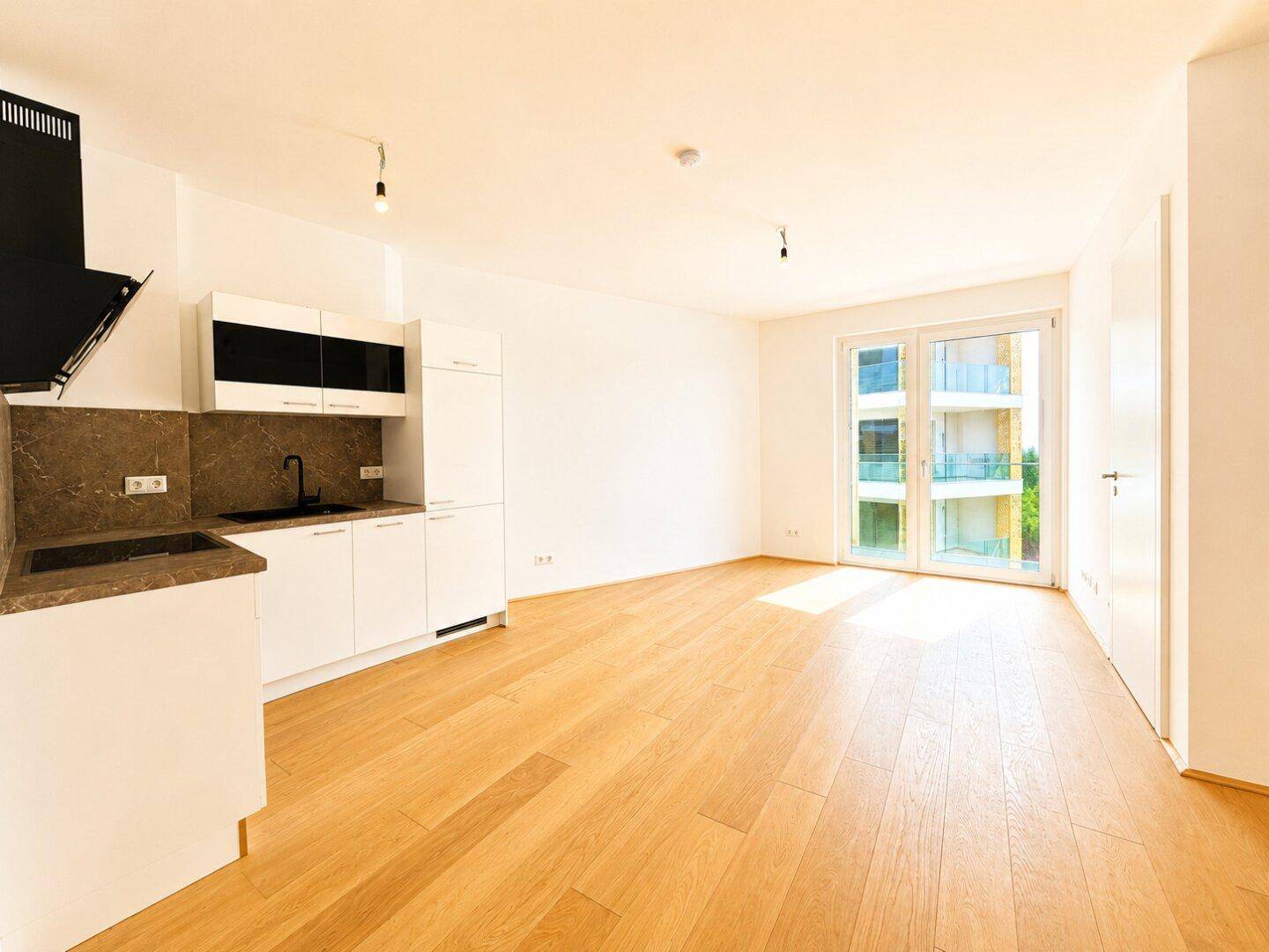 Lichtdurchfluteter Wohnraum mit moderner Küchenzeile, Parkettboden und Zugang zum Balkon.