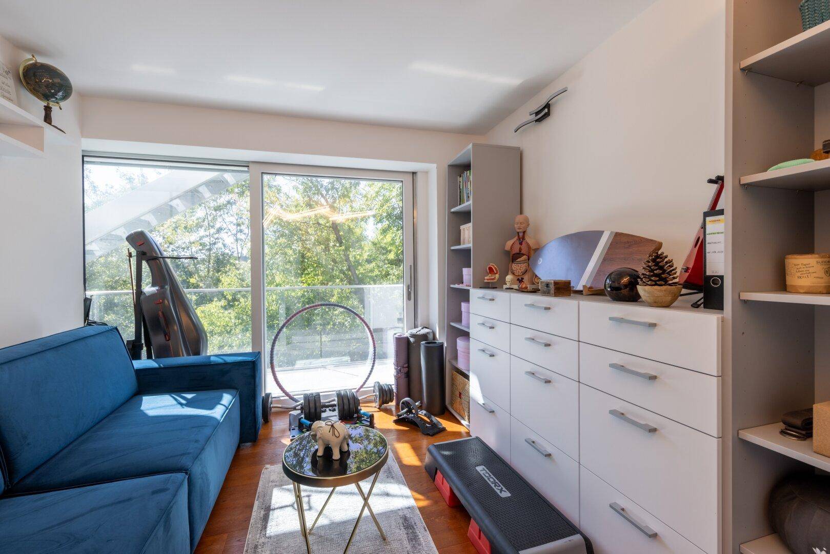 Helles Zimmer mit blauem Sofa, großem Fenster und vielseitiger Nutzung als Hobby- oder Gästezimmer.