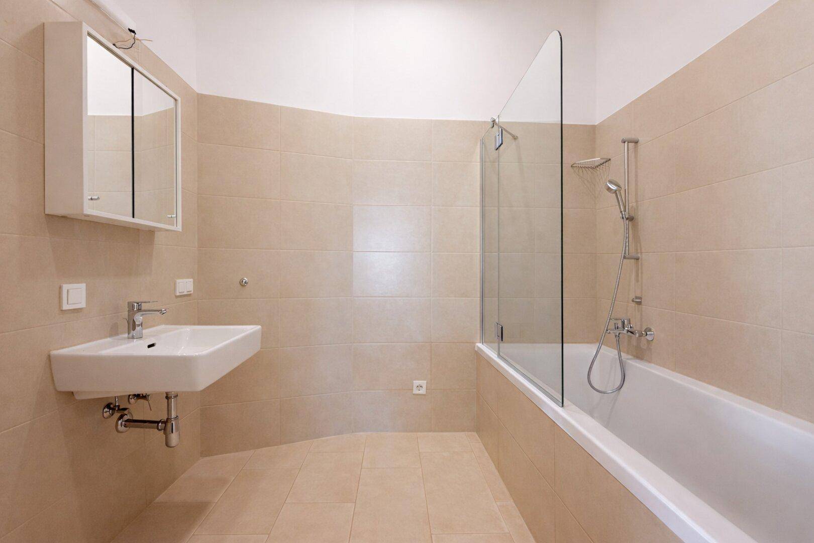 Helles Badezimmer mit beigefarbenen Fliesen, Badewanne mit Duschabtrennung und Waschbecken.