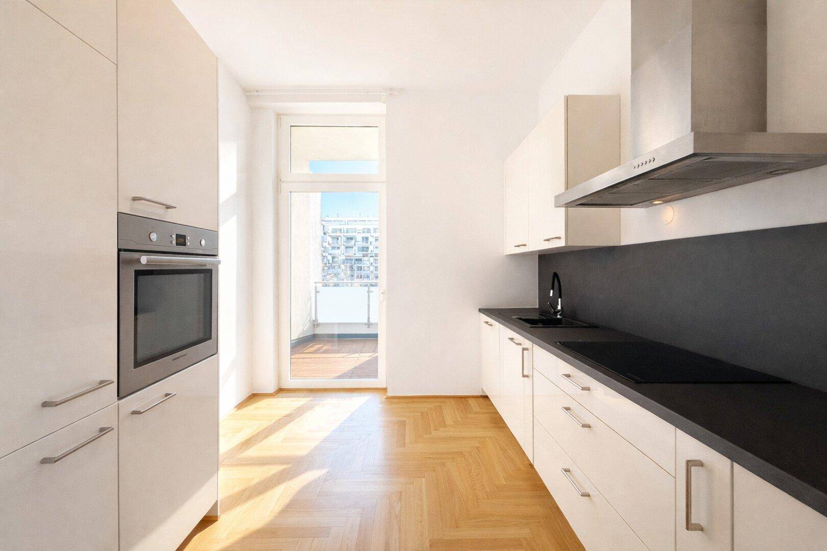 Moderne Küche mit weißen Schränken, dunklen Arbeitsplatten und Zugang zu einem sonnigen Balkon.