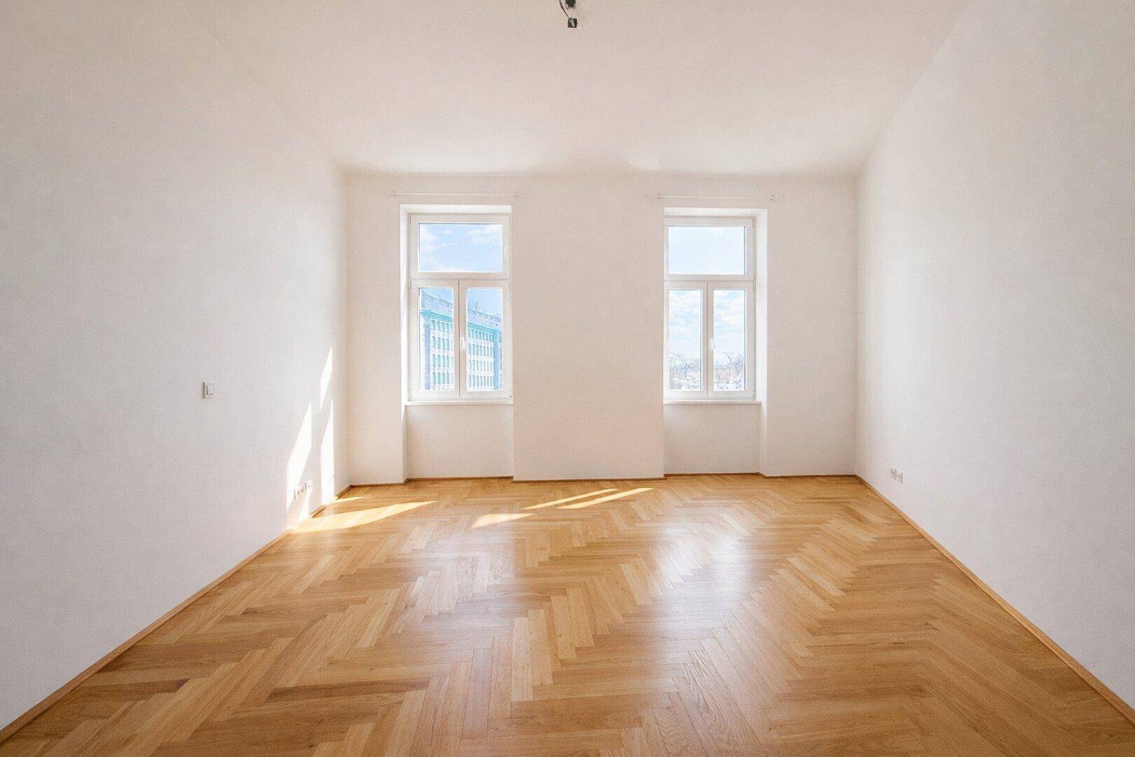 Geräumiges Zimmer mit klassischem Fischgrätparkett und zwei großen Fenstern, die viel Licht hereinlassen.