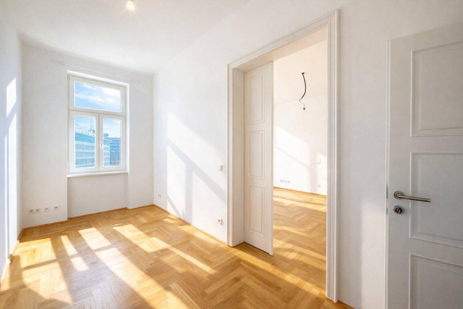 Helles Zimmer mit Fischgrätparkett und einem großen Fenster, das den Raum mit Tageslicht füllt.