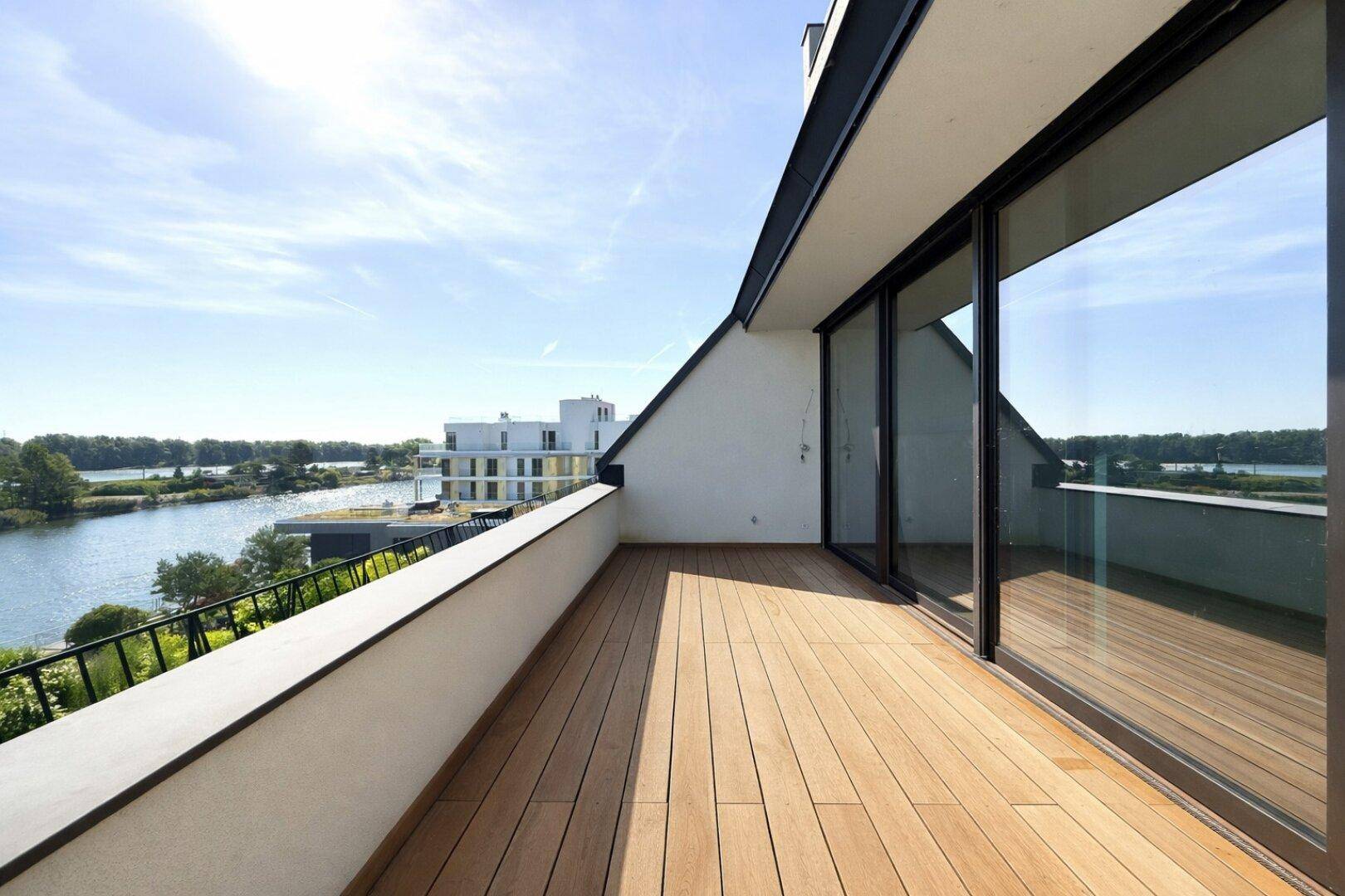 Großer Balkon mit Holzboden und Geländer, der einen weiten Blick auf den See bietet.