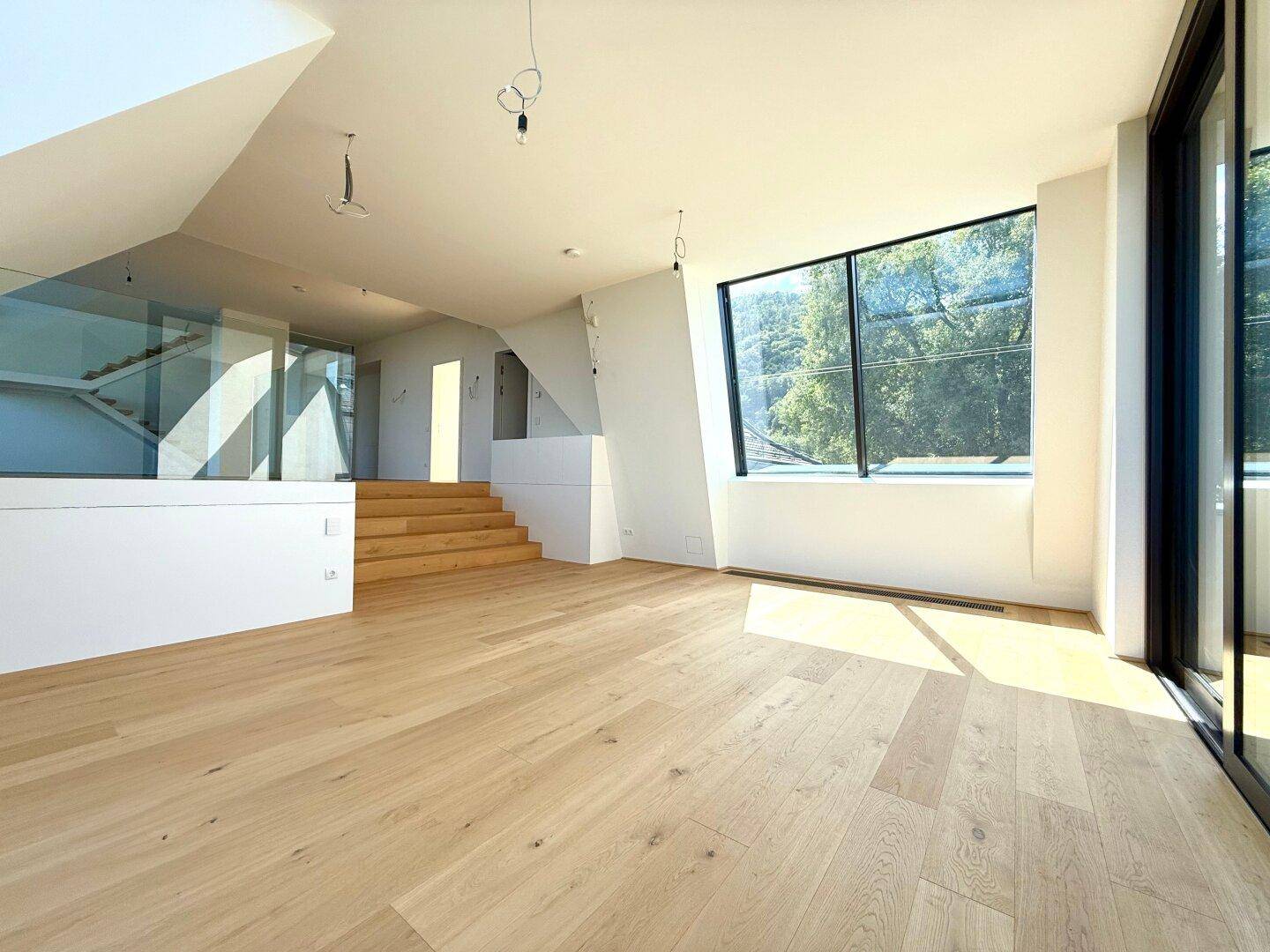 Heller Wohnbereich mit Holzboden, moderner Treppe und großen Fenstern mit Blick ins Grüne.