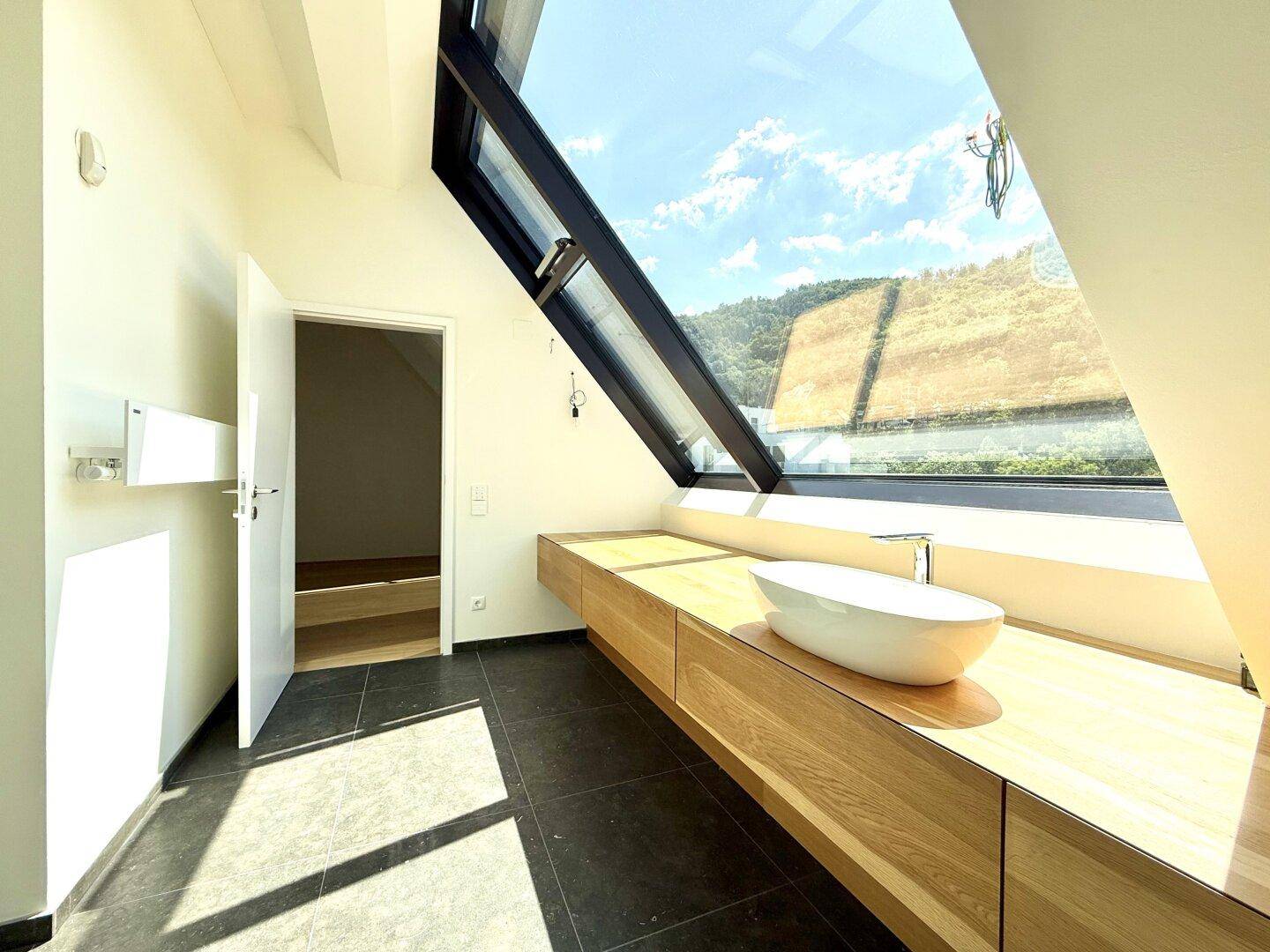 Modernes Badezimmer mit dunklen Fliesen, Holzwaschtisch und großem Dachfenster mit Ausblick.