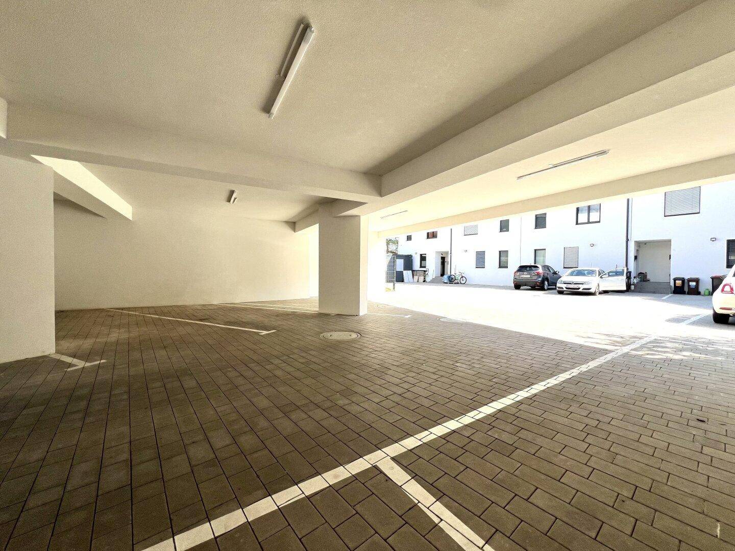 Überdachter Parkbereich mit gepflastertem Boden und markierten Stellplätzen unter dem Gebäude.
