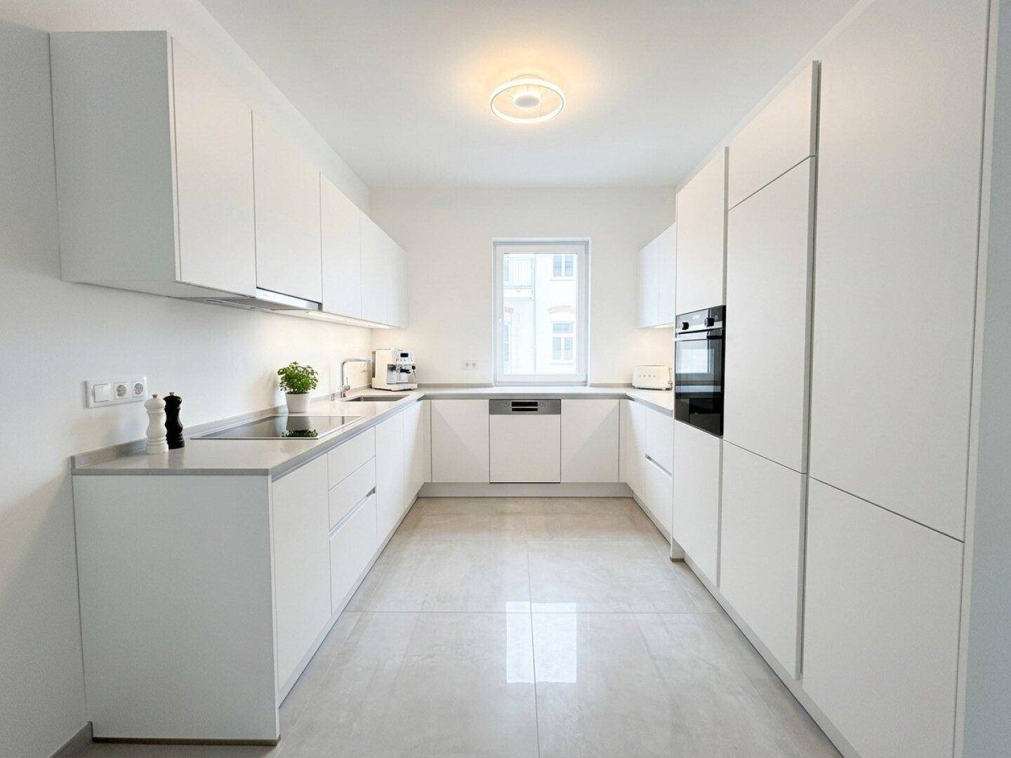 Moderne weiße Küche mit Einbaugeräten, Fliesenboden und einem Fenster für Tageslicht.