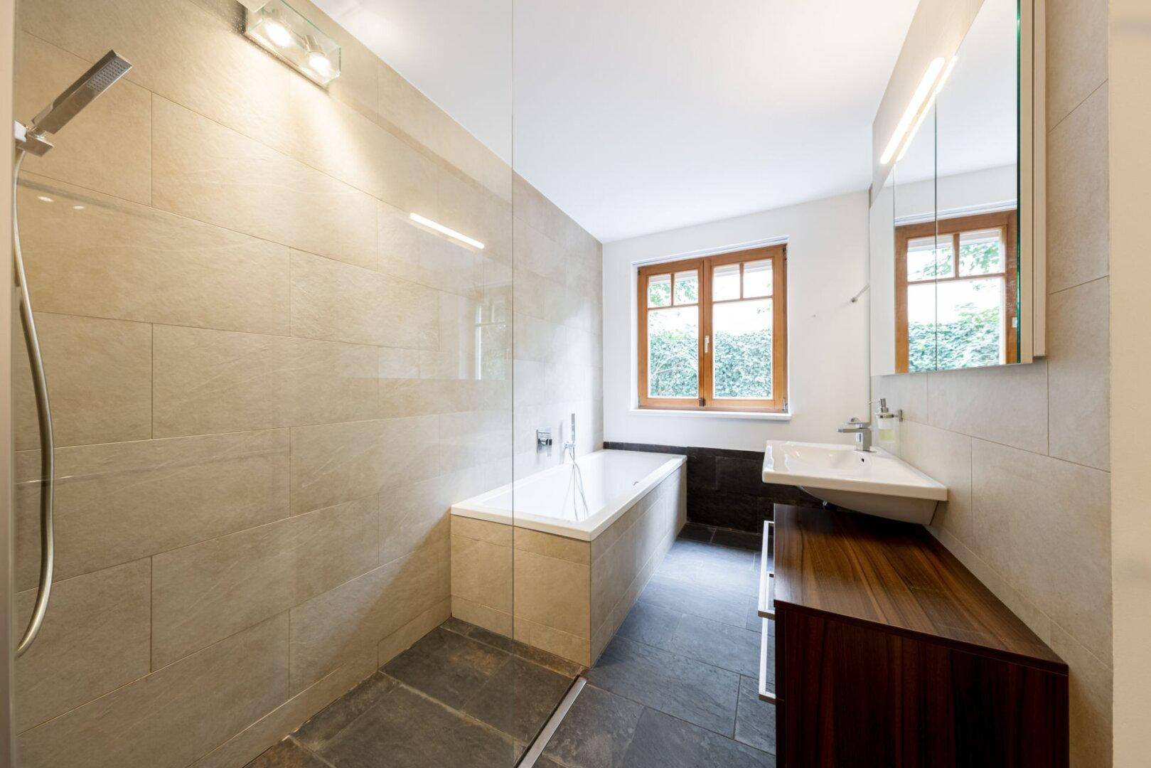 Modernes Badezimmer mit Badewanne, Duschbereich, Einzelwaschbecken und Fenster mit Grünblick.