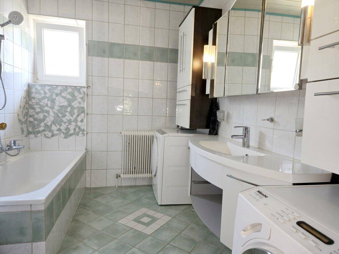 Gefliestes Badezimmer mit Badewanne, Waschbecken, Spiegelschrank und Fenster.