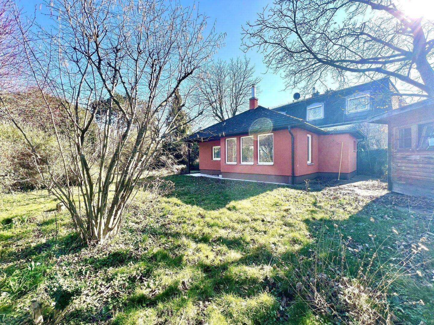 Gartenbereich mit altem Baumbestand und Blick auf ein rotes Haus unter blauem Himmel.