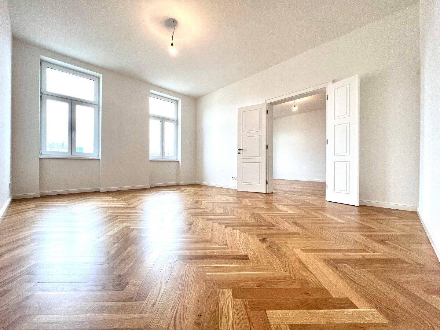 Großes, helles Zimmer mit klassischem Fischgrätparkett und hohen Doppeltüren, die zu einem weiteren Raum führen.