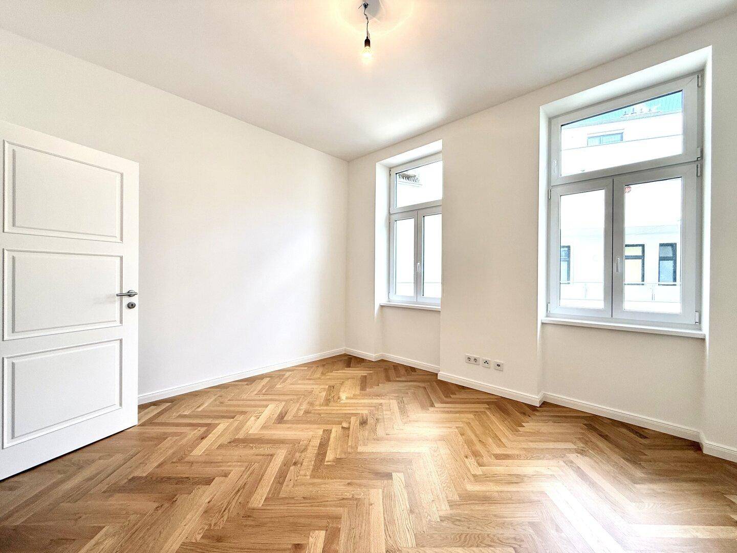 Helles Schlafzimmer mit elegantem Fischgrätparkett und zwei Fenstern, die für viel Tageslicht sorgen.