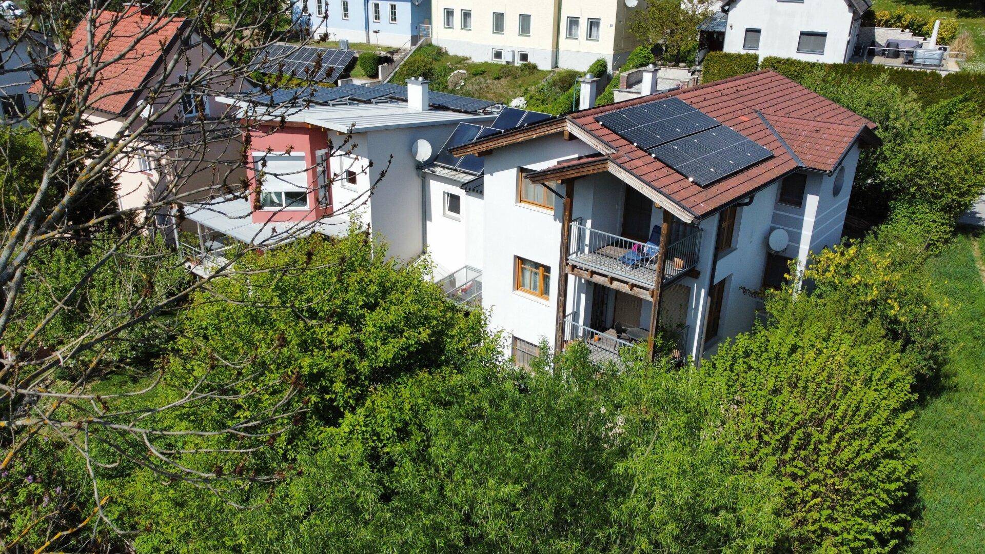 Haus mit mehreren Balkonen und Photovoltaikanlage auf dem Dach, umgeben von üppiger Vegetation.