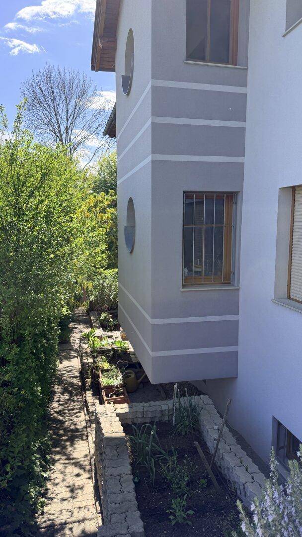 Schmaler Gartenweg entlang der Hausfassade mit Hochbeeten und üppiger grüner Bepflanzung.