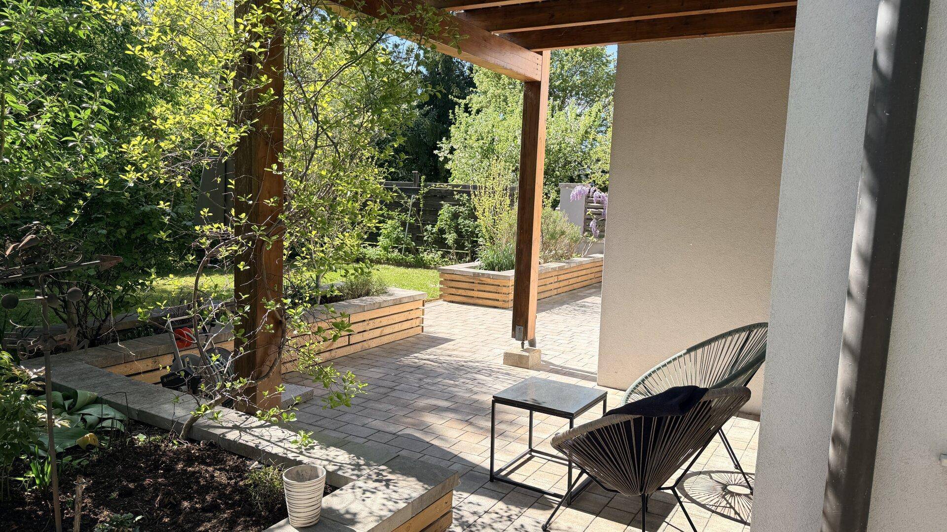 Sonniger Terrassenbereich mit Sitzgelegenheiten unter einer Holzpergola und Blick in den Garten.