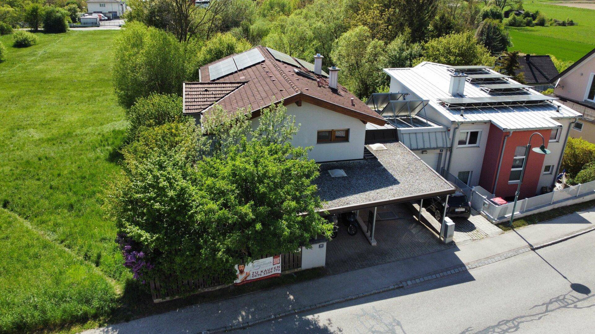 Luftaufnahme eines Hauses mit Photovoltaikanlage und Carport, umgeben von Bäumen und Nachbargebäuden.