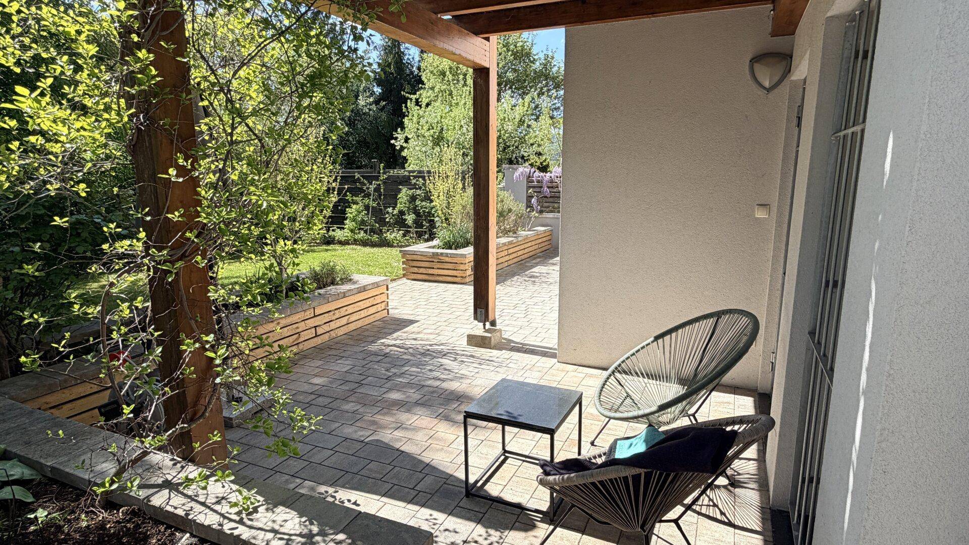 Sonniger Terrassenbereich mit Sitzgelegenheiten unter einer Holzpergola und Blick in den Garten.