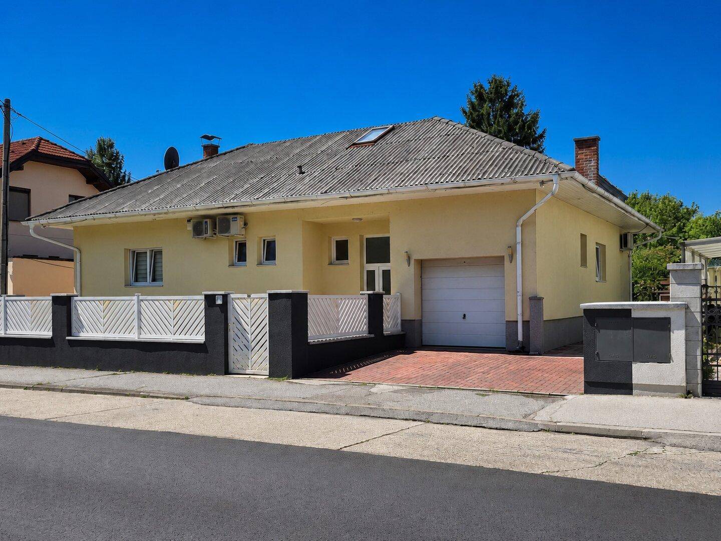 Vorderansicht des Hauses mit Garage, Einfahrt und Zaun unter blauem Himmel.
