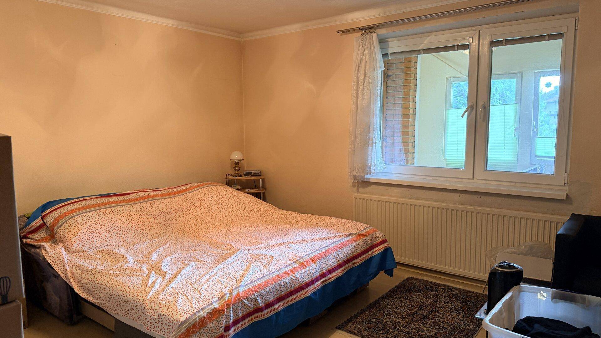 Helles Schlafzimmer mit Doppelbett, Fenster und schlichten Möbeln in warmen Farbtönen.