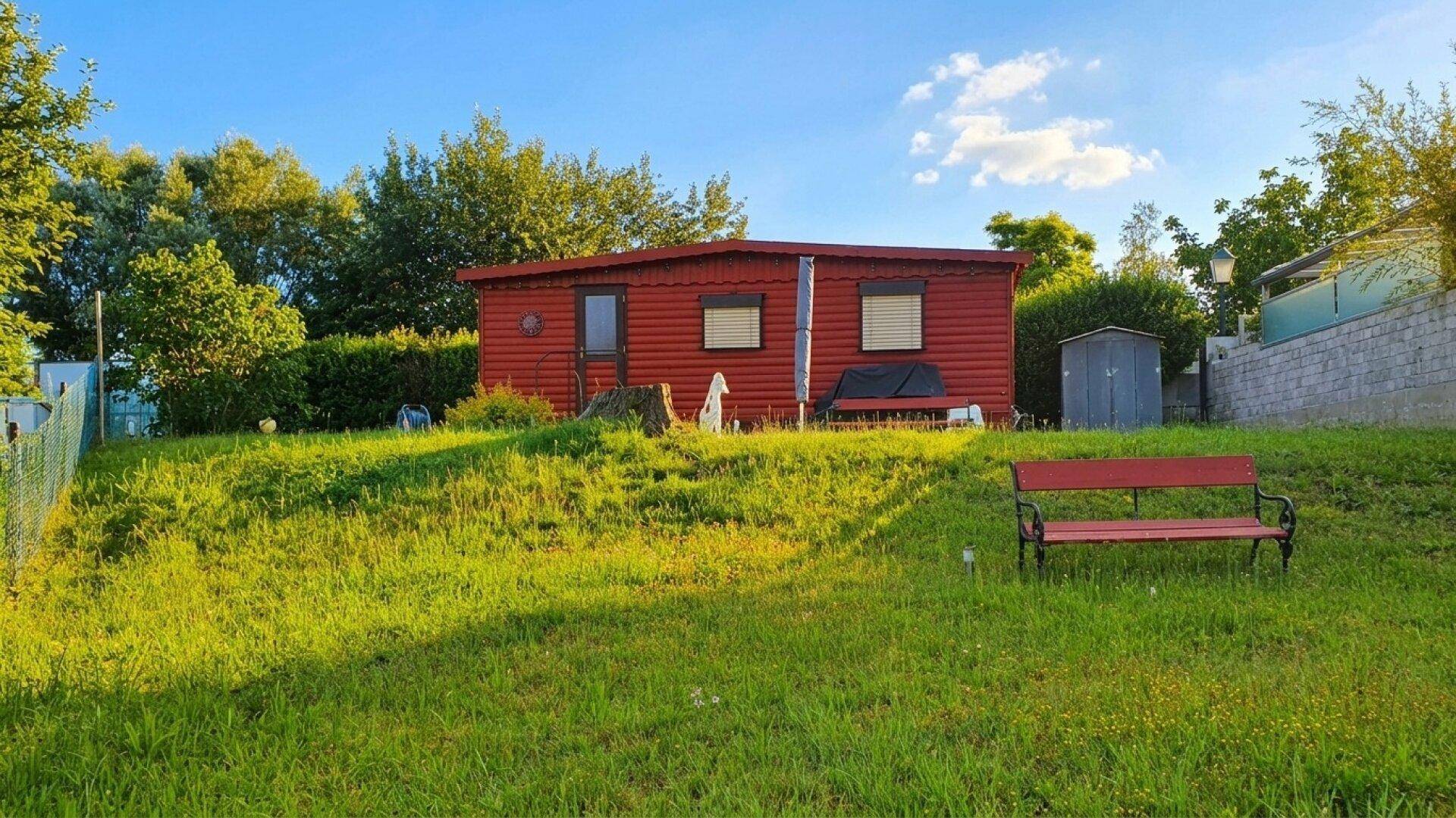 Sonniger Gartenbereich mit einer roten Hütte und einer Holzbank, umgeben von üppigem Grün.
