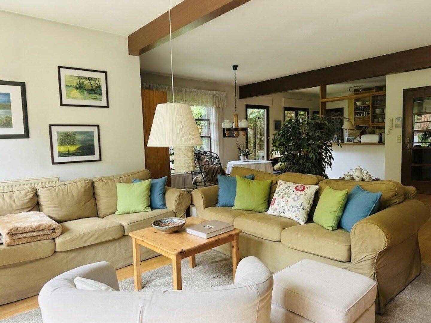 Gemütliches Wohnzimmer mit beigen Sofas, bunten Kissen und Holzbalken, offen zum Essbereich.