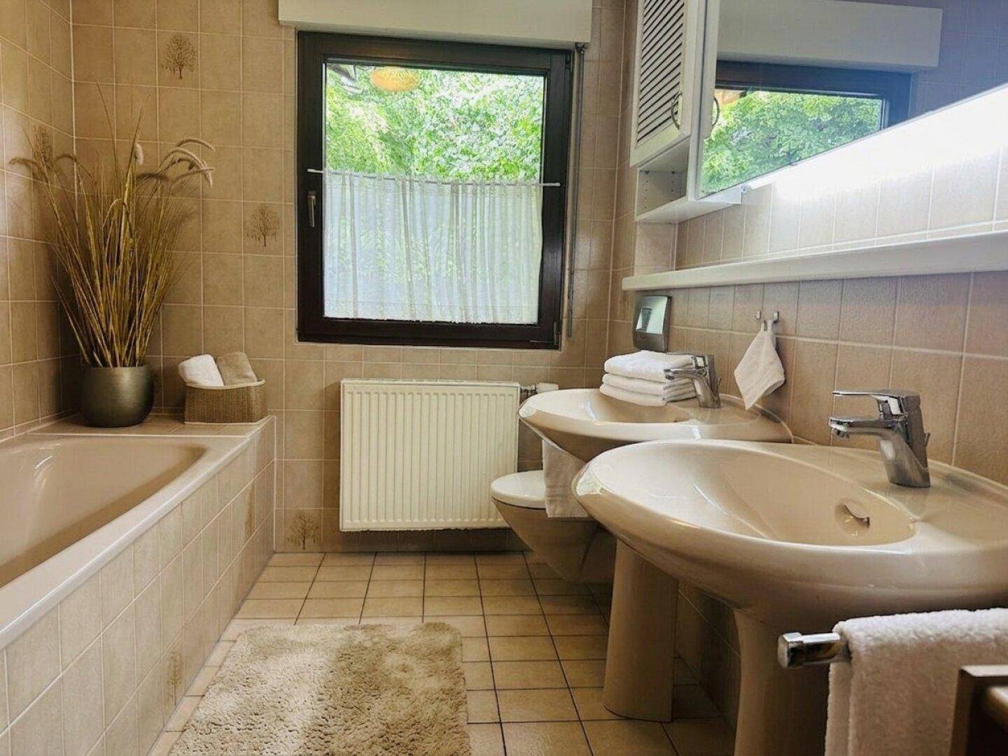 Badezimmer mit beigen Fliesen, Badewanne, Doppelwaschbecken und Fenster mit Blick ins Grüne.