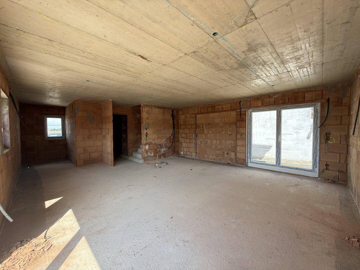 Rohbau eines Innenraums mit unverputzten Ziegelwänden, Betonboden und großen Fensteröffnungen.