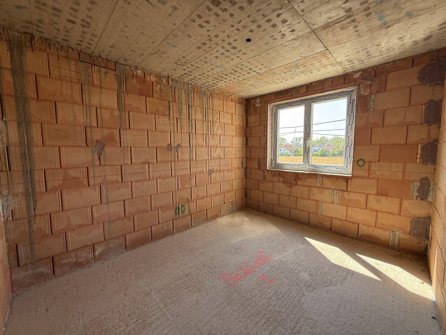 Rohbau eines Zimmers mit unverputzten Ziegelwänden, Betonboden und einem Fenster.