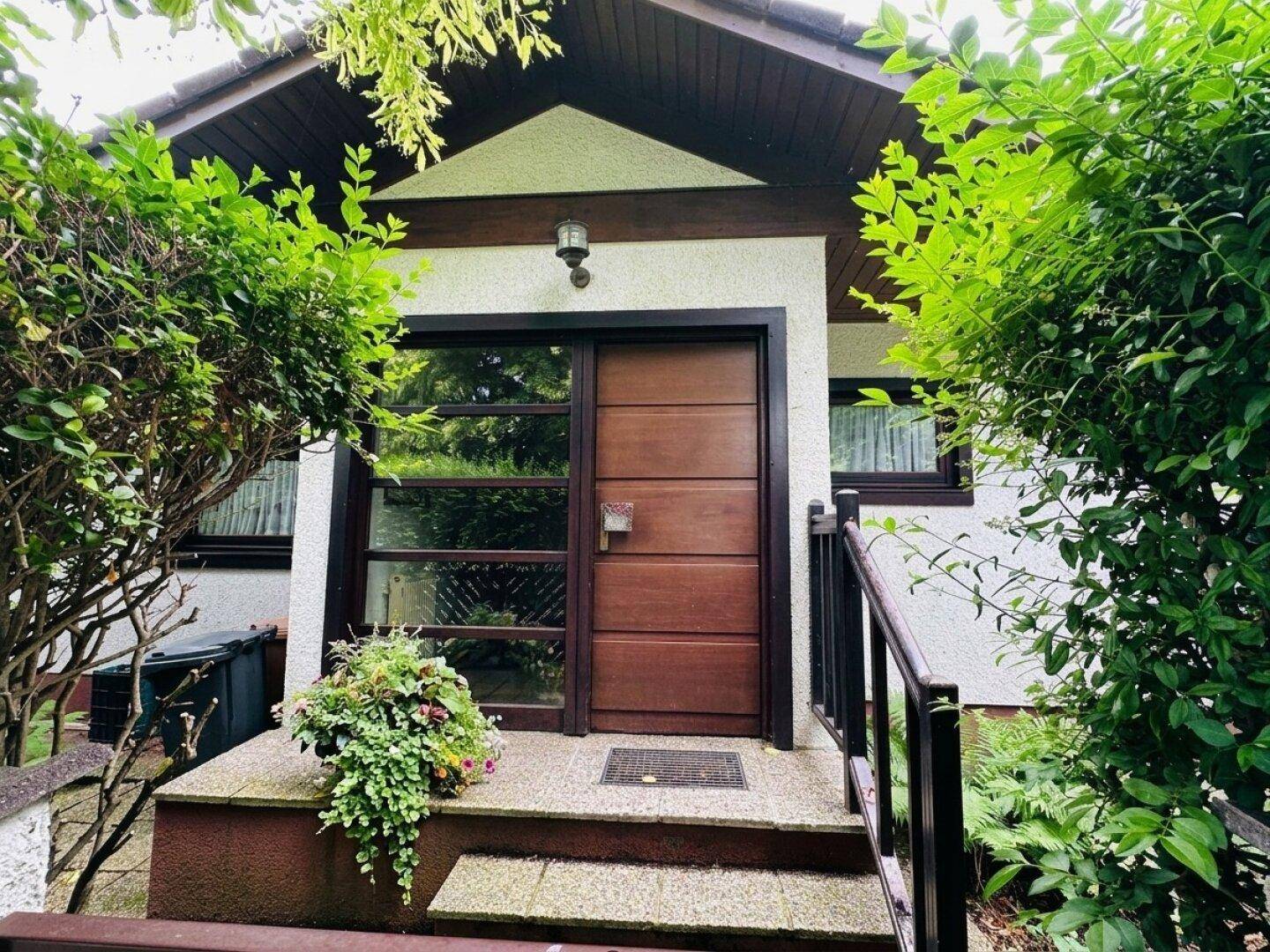 Eingangsbereich des Hauses mit Holztür, Treppe und umgebender Bepflanzung.