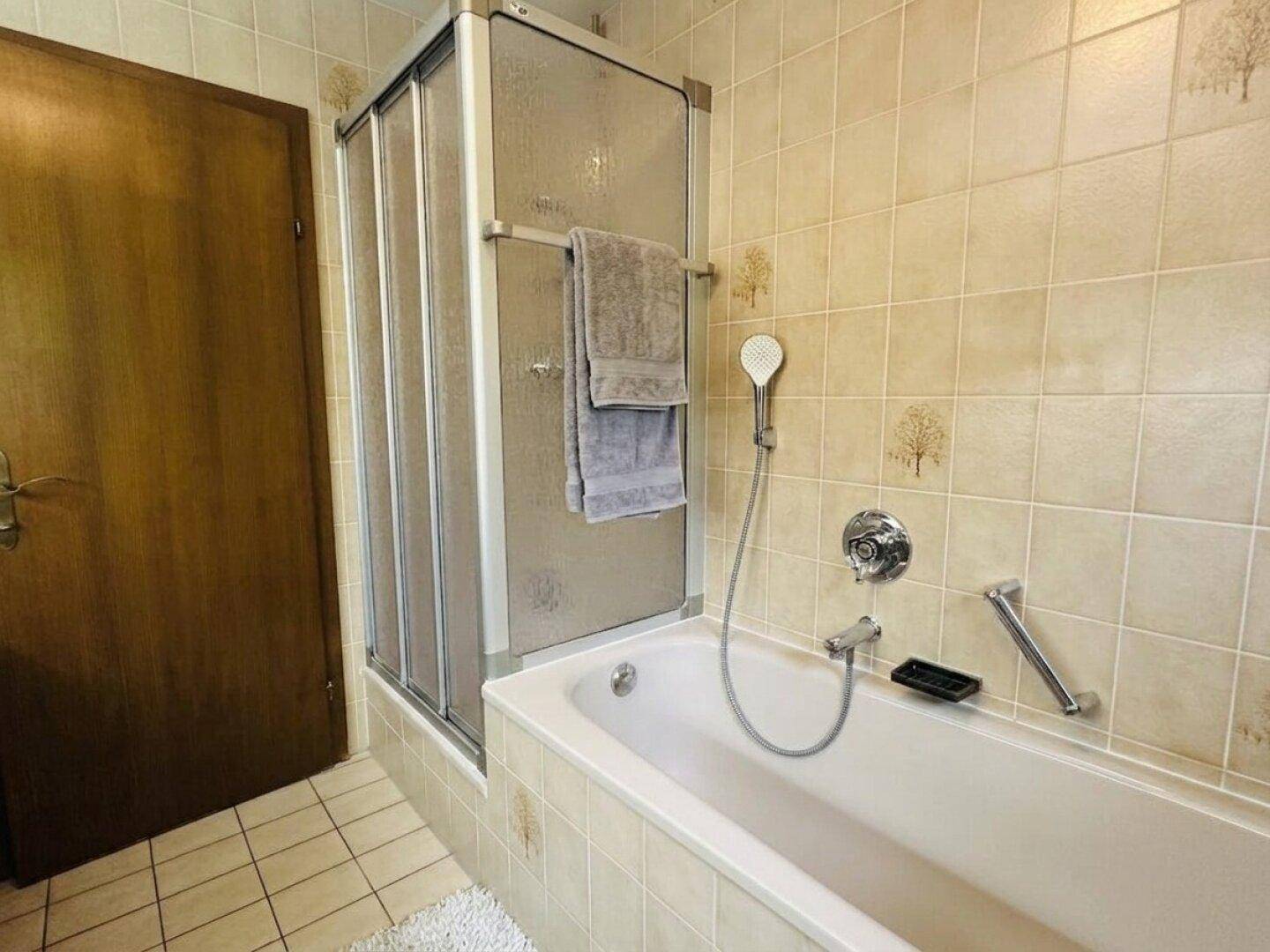 Badezimmer mit beigen Fliesen, Badewanne mit Duschabtrennung und Haltegriffen.