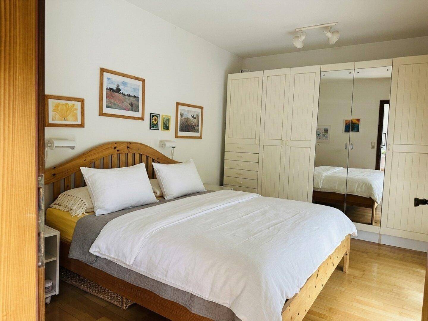 Schlafzimmer mit hellem Holzbett, weißem Kleiderschrank mit Spiegeltüren und Parkettboden.
