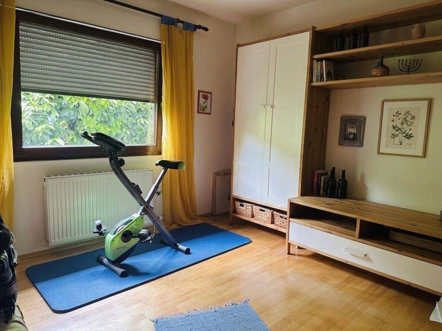 Zimmer eingerichtet als Fitnessraum mit Heimtrainer, Yogamatte und Holzmöbeln.