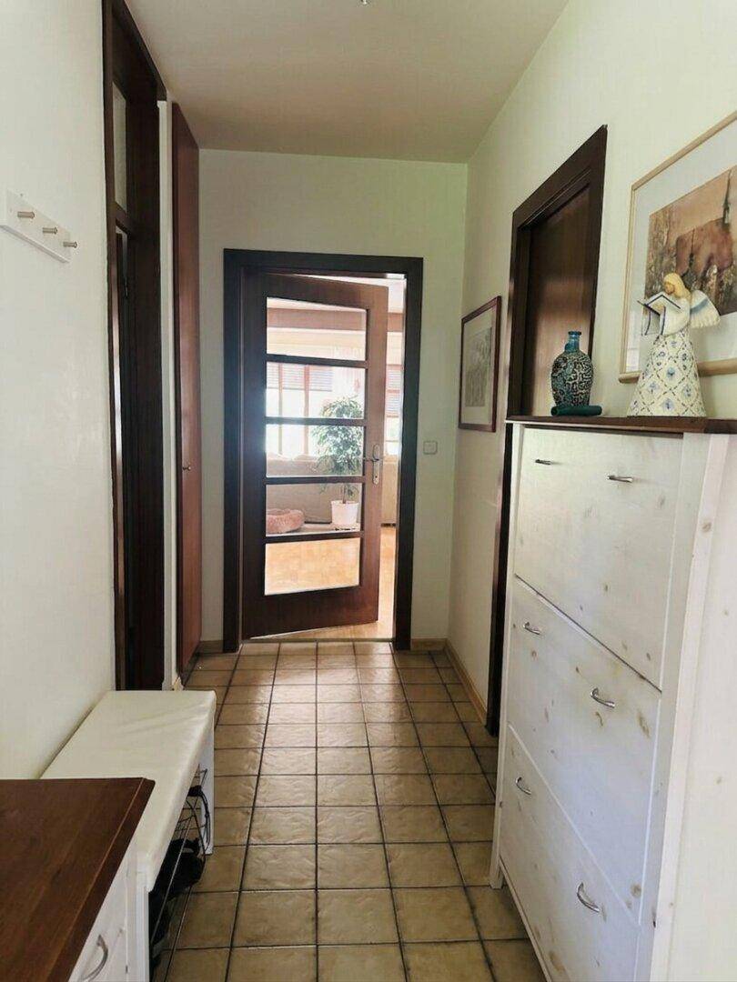 Flur mit Fliesenboden, Schuhschrank und Blick durch eine offene Tür in einen weiteren Raum.