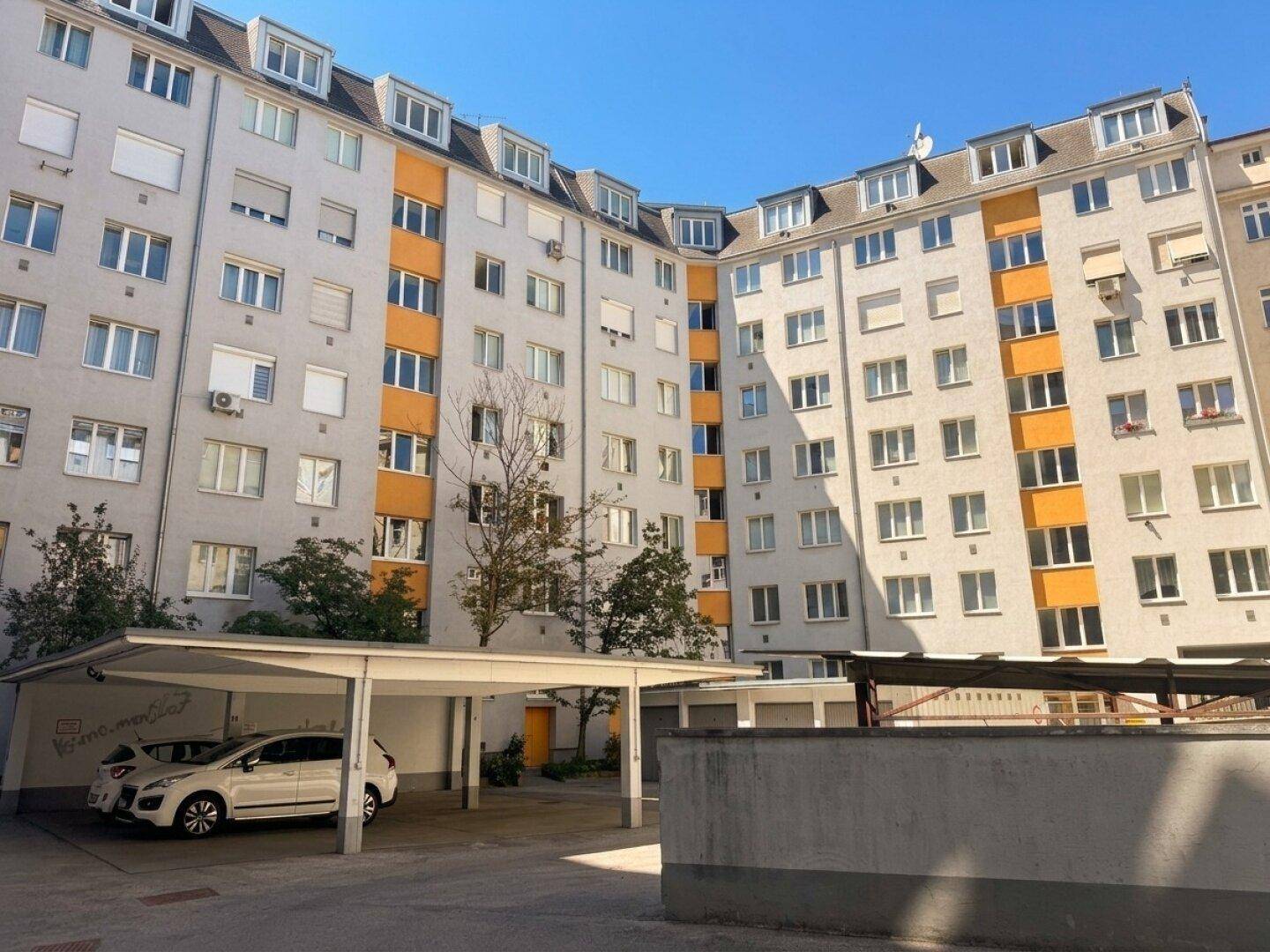 Mehrfamilienhaus mit vielen Fenstern, Balkonen und einem Carport im Vordergrund.