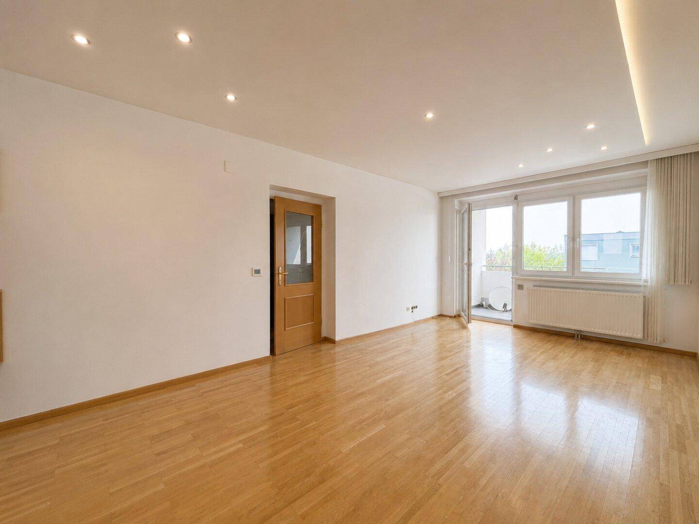Helles Wohnzimmer mit Parkettboden, Einbaustrahlern und Zugang zu einem kleinen Balkon.