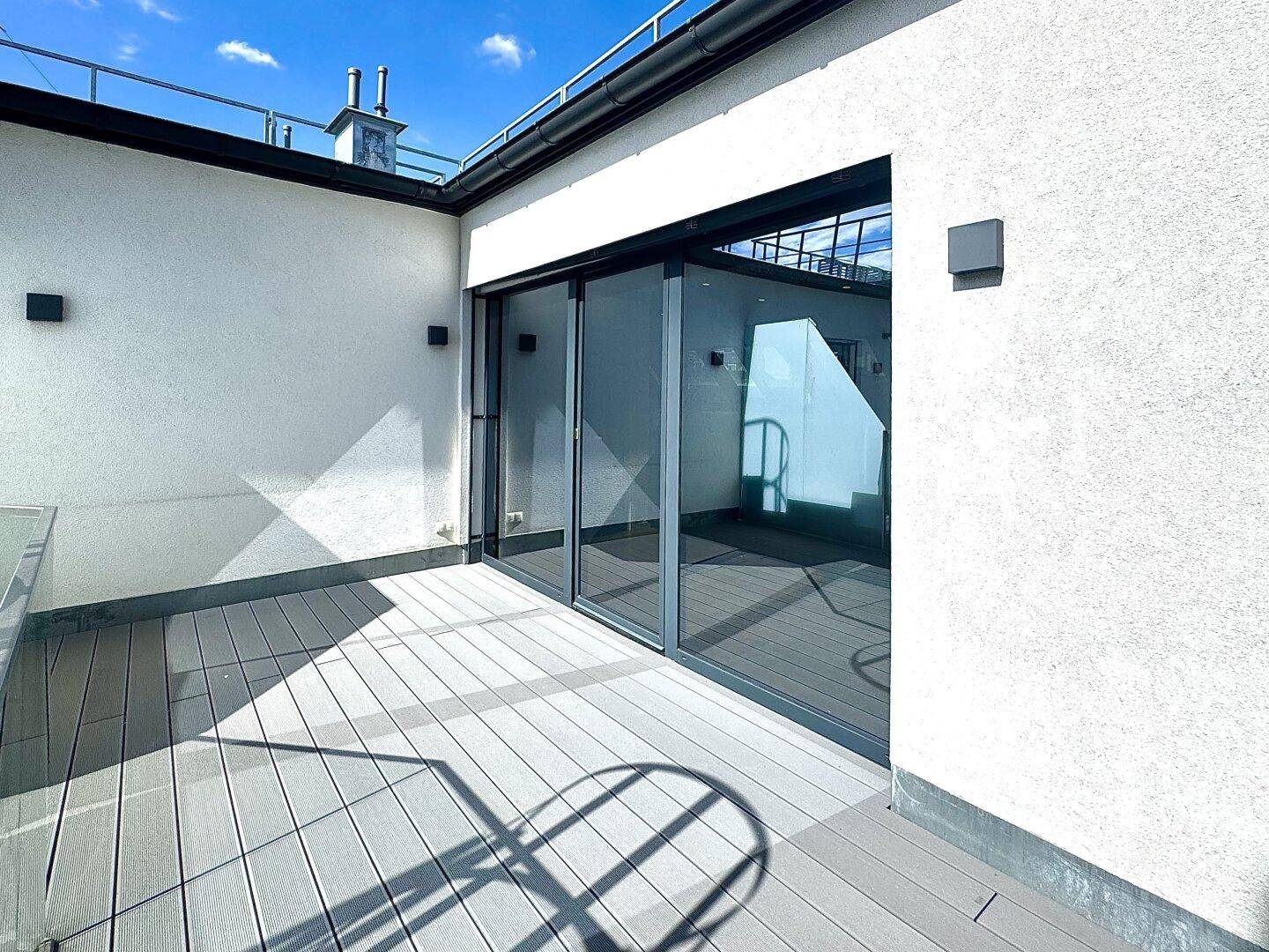 Sonnige Dachterrasse mit Holzdielen und großen Schiebetüren zum Innenbereich.