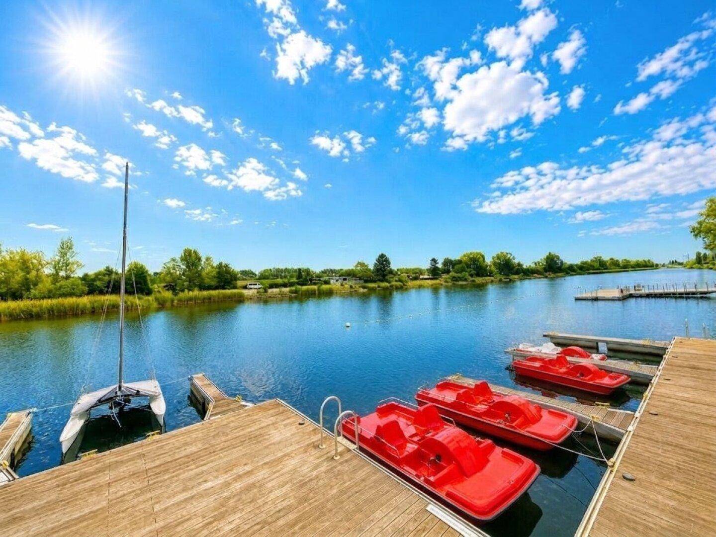 Sonniger Tag am See mit Holzsteg und roten Tretbooten, umgeben von grüner Natur und klarem blauen Himmel.