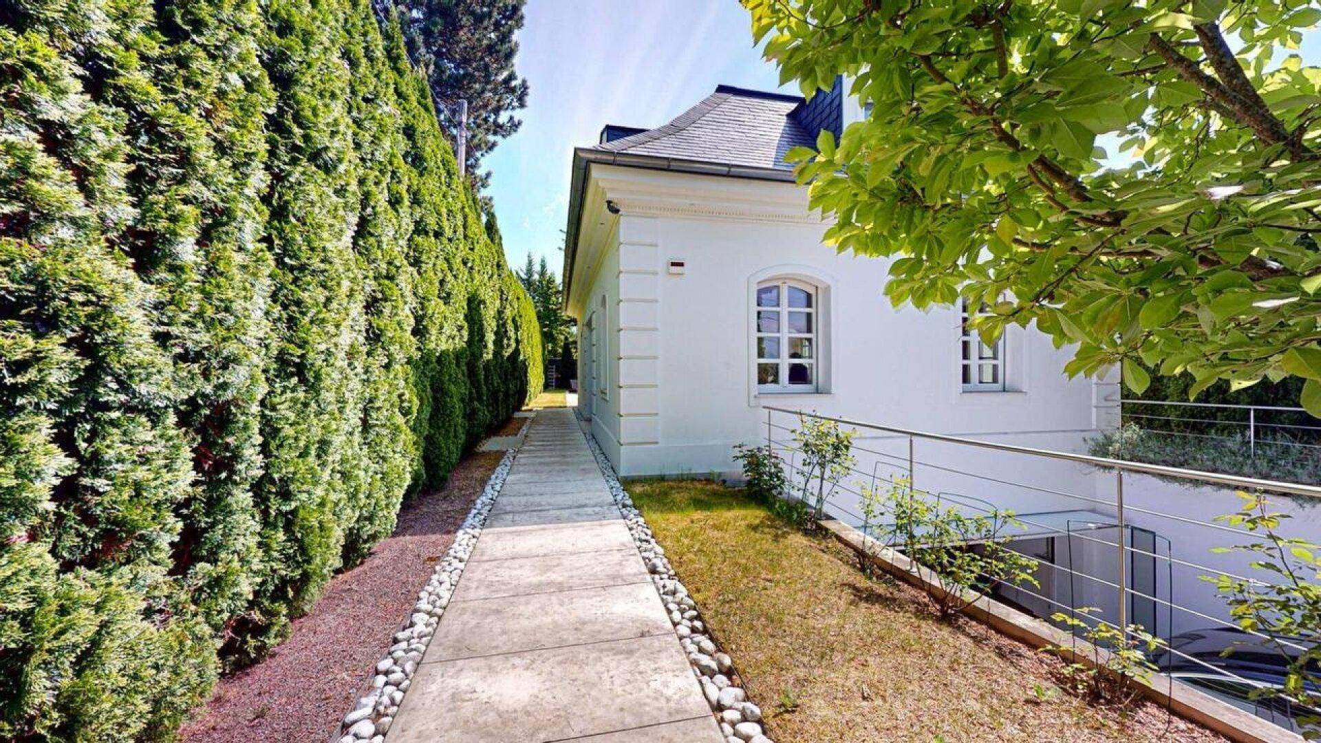 Seitenansicht des Hauses mit gepflegtem Weg, grüner Hecke und Zugang zum Untergeschoss.