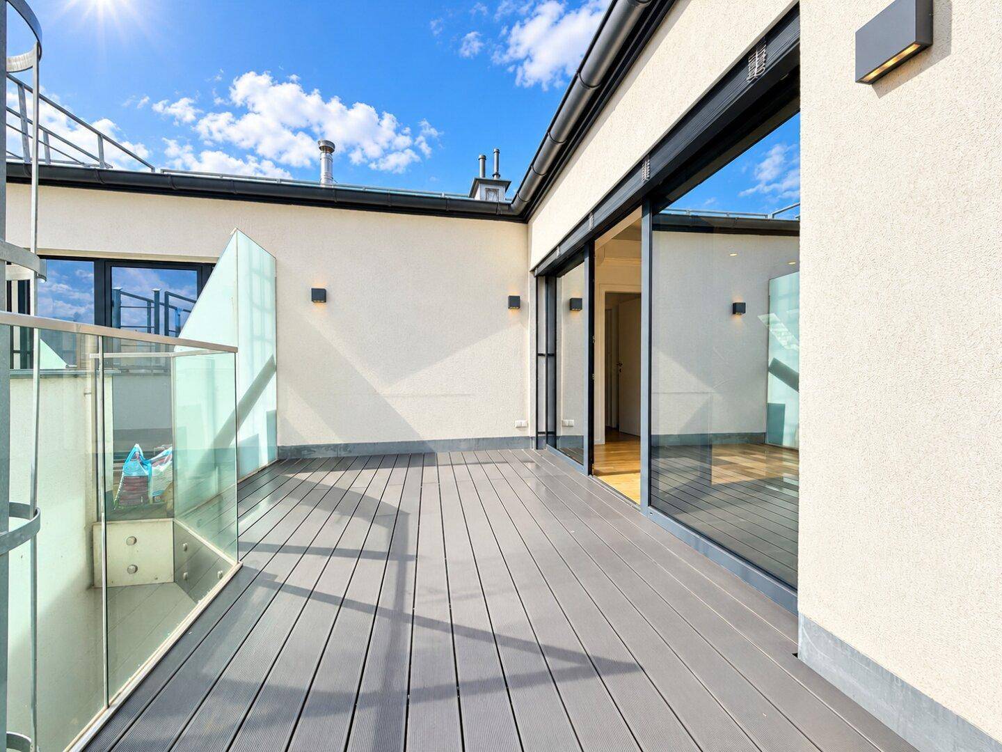 Moderne Dachterrasse mit grauen Dielen, Glasgeländer und Zugang zu den Innenräumen.