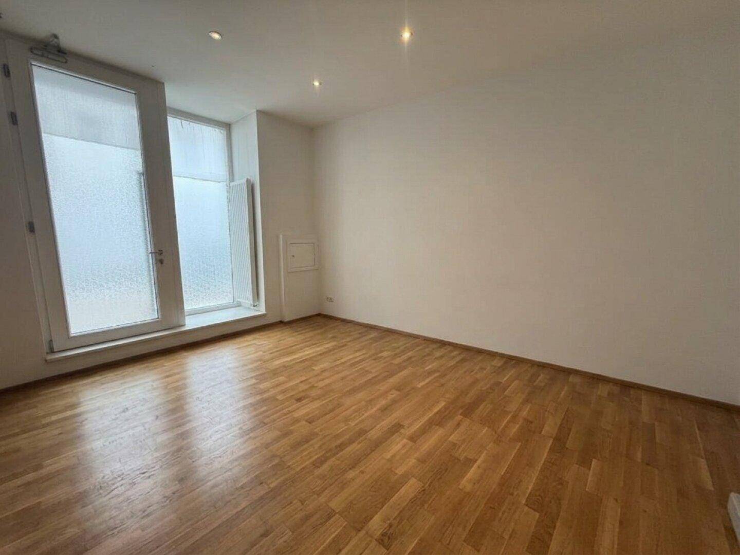Geräumiger, leerer Raum mit Holzboden und hohen Fenstern, die für Helligkeit sorgen.